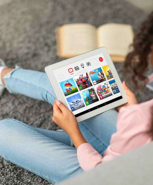 A child sits on the floor of their house against a couch while using YouTube Kids on their tablet